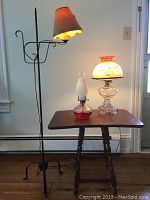 Photo showing all three lamps on a wooden table: black metal floor lamp with red lampshade, oil lamp with red glass base and frosted chimney, and decorative glass globe lamp with floral shade and crack.