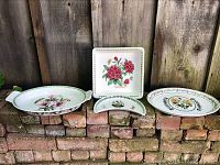 Wide shot showing four Portmeirion pieces on brick wall: oval dish with pink flowers, square tray with red peony and butterflies, crescent half-moon dish with flowers and butterflies, and round plate with multicolored flowers and insects.