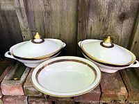 Two lidded serving bowls and one platter displayed together on brick surface, front view.