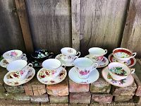 Eight fine bone china tea cups and saucers in various floral patterns displayed on bricks against wood background.