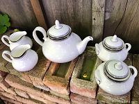 Photo of the full tea set showing teapot, sugar bowl, and two creamers arranged on bricks, highlighting the white porcelain with black detail.