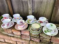 Seven vintage fine bone china teacup and saucer sets displayed on bricks showing floral patterns and brand variety.