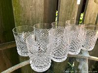 Eight crystal double old fashioned glasses arranged in a row on a glass tabletop against a wood fence.
