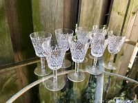 Eight Waterford Crystal Alana stemware glasses arranged in two rows on a glass table outdoors with a wood fence background.