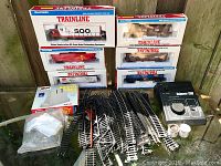 Full lot showing boxed Walthers Trainline locomotives and freight cars, assorted train tracks, bridge piers, and controller on a glass table outdoors.