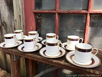 Six white porcelain coffee cups and matching saucers arranged on wooden bench against red window frame