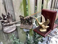 Three vintage bookends placed on glass table outdoors by wooden fence: one flying goose on wooden base, two hunting dogs in bronze metal finish on textured metal bases.