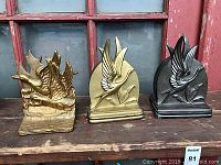 Three cast metal bookends depicting flying geese in different finishes arranged on a wooden surface.