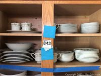 Wide shot of wooden cabinet shelves filled with stacks of white Delux brand Japanese China dishware. Includes plates, bowls, cups, and saucers all with floral trim.