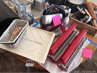 Photo shows collection of kitchenware including the red food wrap dispensers, the white segmented tray, foil pans, glass measuring cups, hand mixer, Sonicare toothbrush, and woven basket with red cloth napkins all arranged on a wooden table.