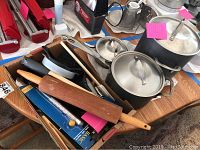 Three Calphalon stainless steel pots with lids and a box of assorted kitchen tools including a rolling pin, knives, melon baller, spatulas, ladle, and other utensils displayed on a table.