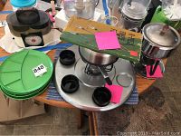 Two fondue pots on metal stands, black fondue cups in a circular tray, large green divided plates stacked nearby, and boxed fondue skewers on table.