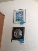 Framed blue and purple abstract floral print and black square wall clock hanging on a white textured wall.