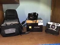 Wide shot of lot showing Nikon COOLPIX P100 camera on original box, vintage Canon camera with attached case, and two black camera bags.