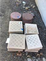 Photo showing stacks of square concrete pavers with lattice pattern and round stepping stones in reddish colors on dirt ground.