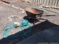 Wheelbarrow, two water cans and garden hose arranged on dirt ground near stone border and fence, showing outdoor setting and condition of items