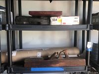 Shelf with a vintage wooden bench plane tool, a cardboard shipment tube, and several cassette tape cases including one labeled 'The New Testament'.