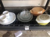 Overview showing 8 clear plates, clear etched glass cup and saucer, 2 cream-colored floral mugs with large matching plates, 2 Pyrex casseroles: yellow with floral lid and amber with lid.