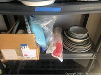 Overall view of the lot including cardboard box with disposable items, plastic wrapped disposable cups, blue disposable tablecloths, and stack of stoneware dishes on shelf.