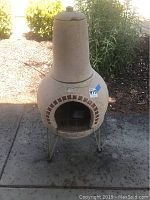 Full view of beige outdoor fire chimney standing on metal legs outside on concrete with garden background.