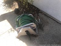 Green garden hose wound on a white plastic hose reel sitting on concrete next to a wall outdoors.