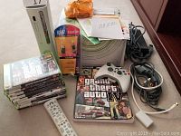 Overview of Xbox 360 console, controller, remote, cords, games, and boxed USB2 cable showing lot contents