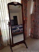 Wood and glass tilting full-length mirror standing upright next to window with curtains, showing overall view and room reflection