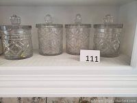Four heavy round crystal canisters with lids arranged on a white shelf, showing their octagonal knob handles and cut glass design.
