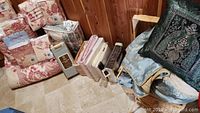 Photo showing an array of packaged and some opened bed linens, mostly patterned in red and cream, with some folded linens in blue and pink on a wooden floor.