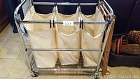 Front view of the laundry cart showing three cream-colored removable canvas bags on stainless steel frame with wheels.