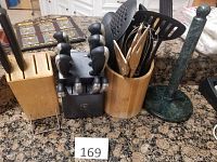 Photo showing JA Henckels Everedge Plus black knife block with 6 knives, bamboo utensil holder with several cooking utensils, empty wooden knife block, and green granite paper towel holder on granite countertop.
