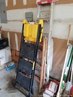 Photo of three ladders leaning against garage wall, showing black step ladder with yellow top, wooden ladder, and aluminum extension ladder.