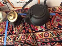 Photo showing cast iron kettle, Ron fire starter with brass lid, and iron dinner chimes on patterned rug.
