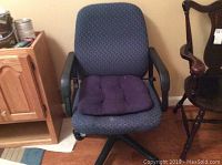 Full view of the dark blue fabric office chair with black armrests and wheeled black base against a wall near wooden furniture.