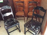 Four chairs including two vintage black wood chairs with paint splatter and two antique wood chairs with recaned cane seats, positioned against a wooden paneled wall.