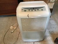 Front view of white Maytag dehumidifier showing ventilation grille and cord on carpet, with minor paint damage on front lower area.