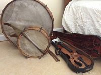Two antique wooden frame drums with torn skins and drumsticks next to a vintage violin in open case with bow.