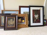 Seven pieces of wall art resting on carpeted floor showing variety of frames and art types including botanical leaf prints, still life painting, and signed Native American piece.