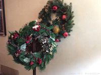 Green holiday wreath with red flower and berry decorations, and other wreath in background with red ornaments and pine cones.