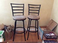 Pair of bar stools showing full view including legs and seats with worn faux suede covers, placed against a wall with cardboard boxes nearby.