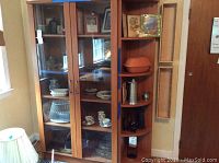 Photo of a wooden bookcase with glass framed doors and corner open shelves showing various decorative items inside and on the shelves.