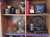 Cupboard shelf showing silver plated platters and bowls, pressed glass dishes, and small decorative framed items