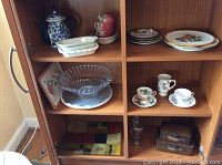 Shelved collection showing blue and white pitcher, ceramic dishes, floral cups and saucers, two miniature leather suitcases, and glass bakeware
