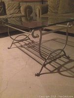 Side angle image of the coffee table showing the silver metal frame with curved scrollwork and the clear glass top, placed on carpeted floor.