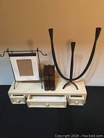 Front view of all five items on display: white wooden wall shelf with three drawers, two brown wooden book boxes side by side, black wrought iron candle holder, and metal picture frame stand.