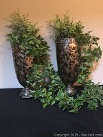 Two metal wall vases with faux greenery on a black surface against beige wall.
