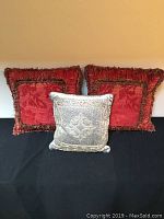 Front view of three cushions: two large red cushions with floral patterns and fringed edges, and one smaller beige cushion with intricate design.