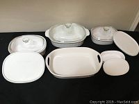 Overhead view of assortment of white CorningWare casserole dishes with a mix of glass and plastic lids on black cloth surface.
