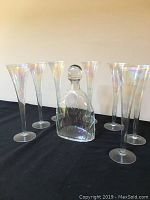 Front view of the decanter with stopper surrounded by six champagne flutes showing iridescent finish.