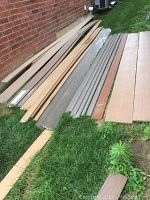Multiple full-length composite and PVC boards laid on grass alongside brick wall
